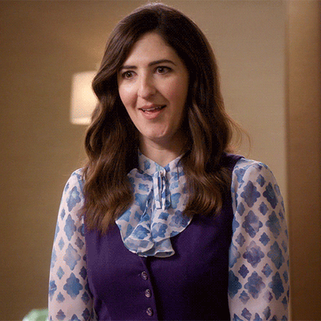 A woman with wavy brown hair, wearing a patterned blouse with a ruffled collar and a purple vest, is smiling.