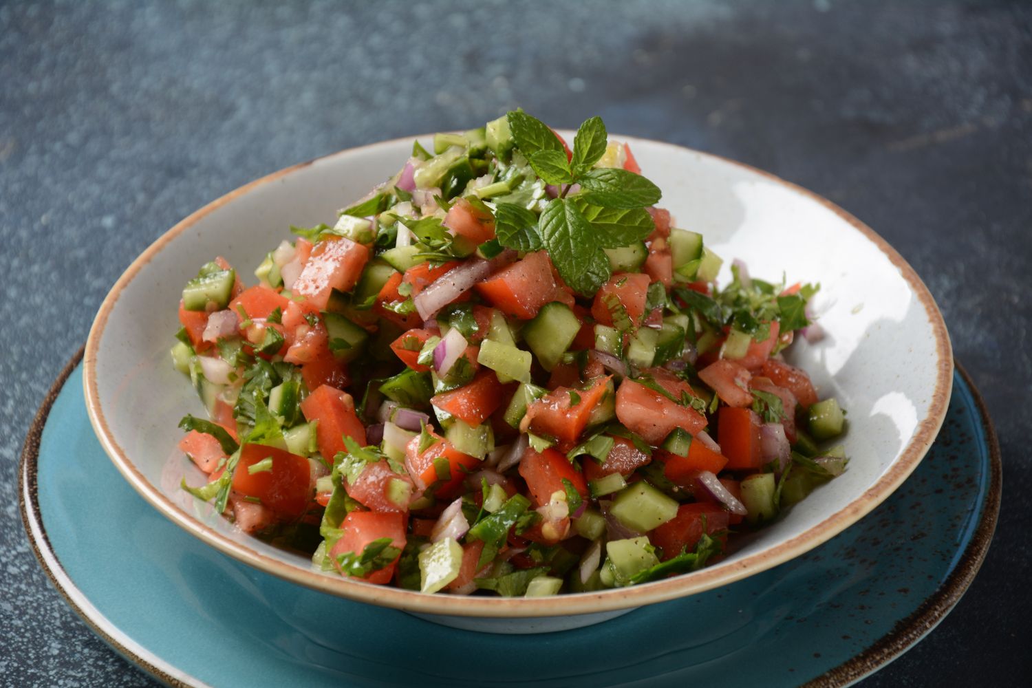 A bowl of fresh vegetable salad containing diced tomatoes, cucumbers, onions, and herbs, garnished with a sprig of mint. The bowl is placed on a teal plate against a dark background.