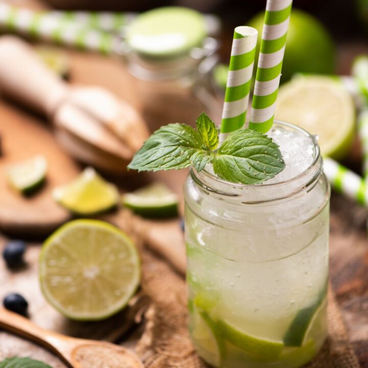 A glass jar filled with a lime drink, ice, and lime slices, garnished with mint leaves and two green striped straws. The drink is placed on a wooden table with lime wedges around it.